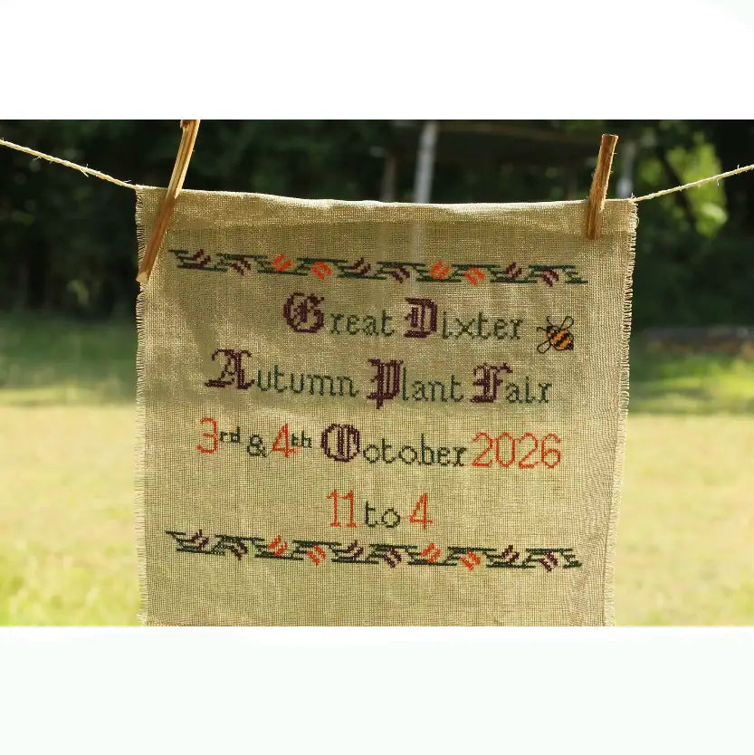 A square beige burlap sign with embroidered text reading 'Great Dixter Autumn Plant Fair 3rd & 4th October 2026 11 to 4' hangs outdoors between two wooden clothespins on a string, with a blurred green garden background.